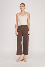 A woman modeling a light beige camisole top with delicate eyelash lace trim at the neckline, paired with brown pants, against a plain light gray studio background.