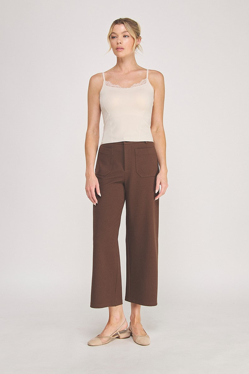 A woman modeling a light beige camisole top with delicate eyelash lace trim at the neckline, paired with brown pants, against a plain light gray studio background.