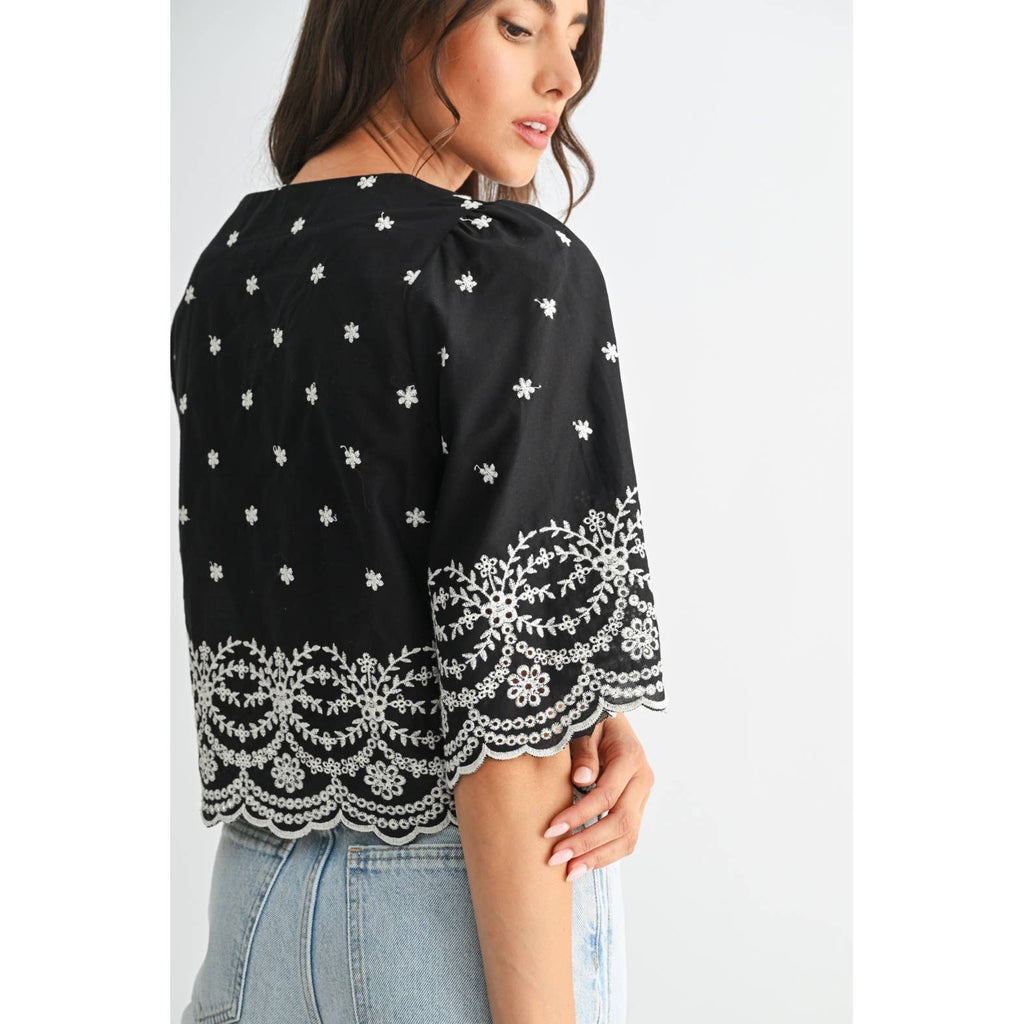 Back view of a woman wearing a black top with white embroidery on a plain background