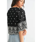 Back view of a woman wearing a black top with white embroidery on a plain background