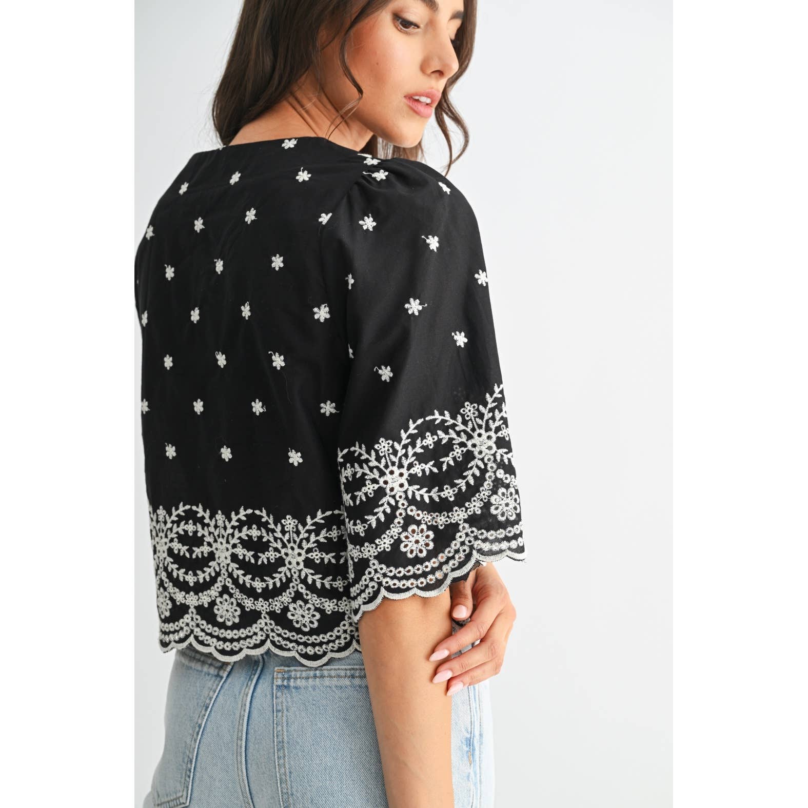Back view of a woman wearing a black top with white embroidery on a plain background