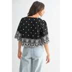 Back viewof a woman wearing a black top with white embroidery on a plain background