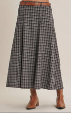 A woman wearing a high-waisted, A-line navy plaid skirt istyled with a brown leather belt and brown ankle boots against a solid beige background.