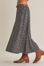 A woman wearing a high-waisted, A-line navy plaid skirt istyled with a brown leather belt and brown ankle boots against a solid beige background.