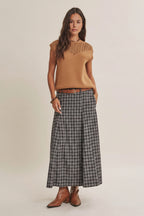A woman modeling a tan sleeveless knit top with a crochet yoke and a navy plaid maxi skirt styled with a brown belt and brown leather boots against a solid beige studio background.