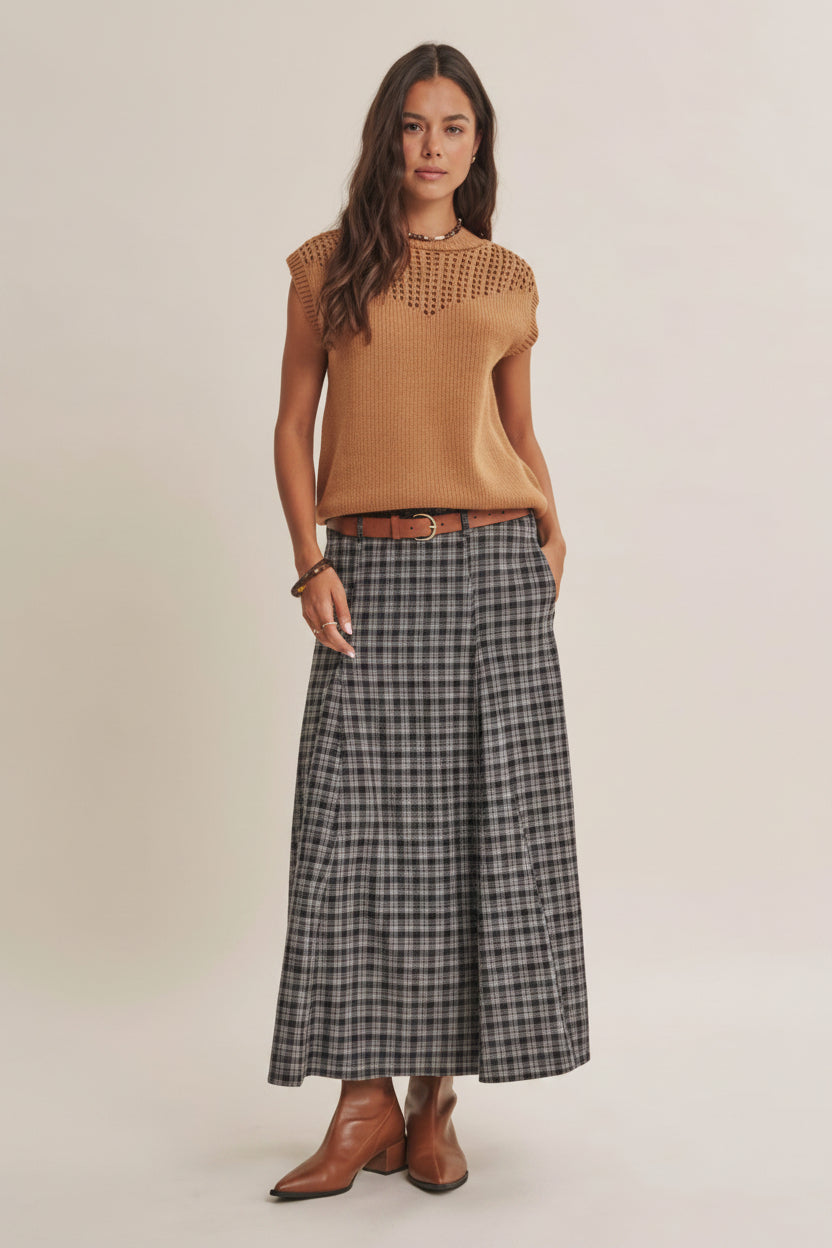 A woman modeling a tan sleeveless knit top with a crochet yoke and a navy plaid maxi skirt styled with a brown belt and brown leather boots against a solid beige studio background.