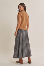 Back view of a woman modeling a tan sleeveless knit top with a crochet yoke and a navy plaid maxi skirt styled with a brown belt and brown leather boots against a solid beige studio background.