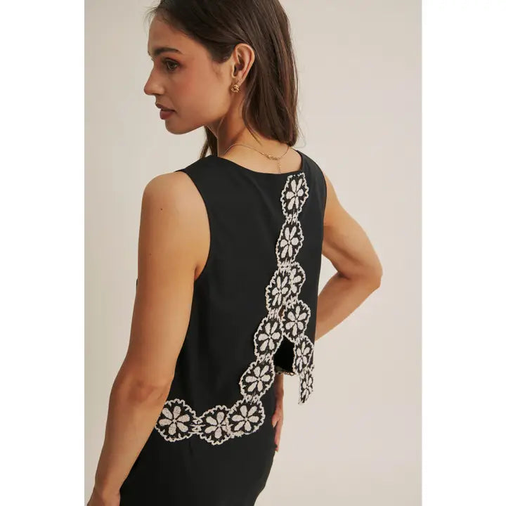 Back view of a woman wearing a black top and skort  with white floral crochet trim on a beige background