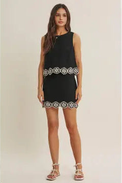Woman wearing a black top and skort  with white floral crochet trim on a beige background