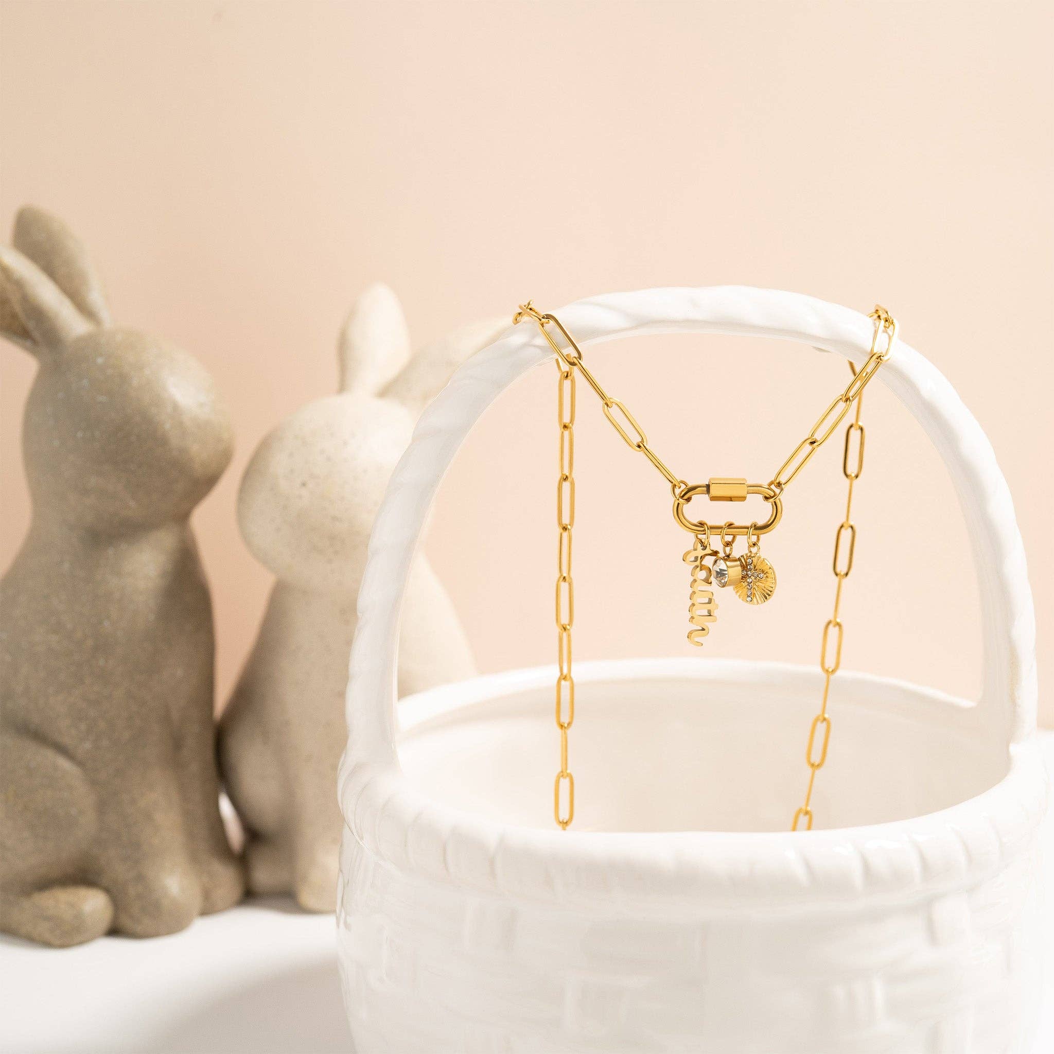 Paperclip carabiner gold chain necklace with charms displayed in a white basket, with ceramic rabbits in the background.