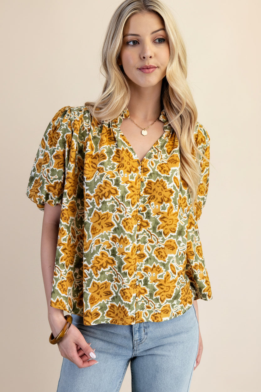 A woman wearing a balloon sleeve blouse with a mustard and green floral pattern, paired with light blue jeans, set against a solid light green background.