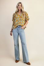 A woman wearing a balloon sleeve blouse with a mustard and green floral pattern, paired with light blue jeans, set against a solid light green background.