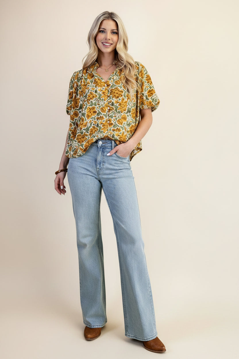 A woman wearing a balloon sleeve blouse with a mustard and green floral pattern, paired with light blue jeans, set against a solid light green background.