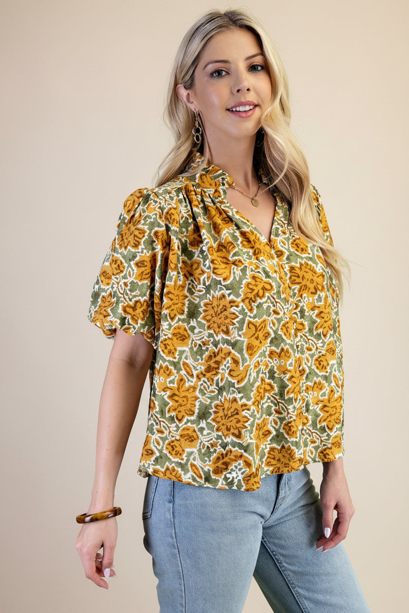A woman wearing a balloon sleeve blouse with a mustard and green floral pattern, paired with light blue jeans, set against a solid light green background.