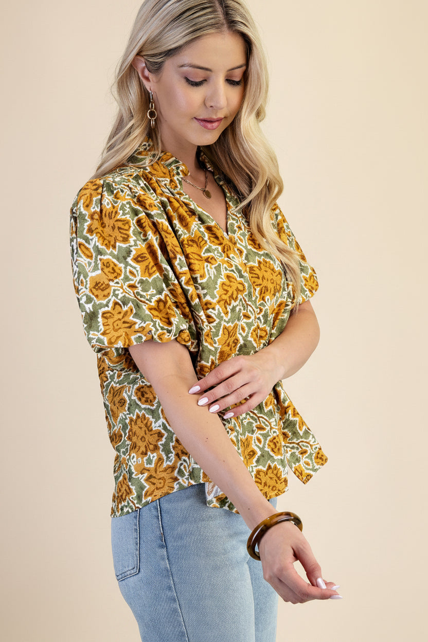 A woman wearing a balloon sleeve blouse with a mustard and green floral pattern, paired with light blue jeans, set against a solid light green background.