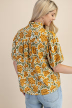 Back view of a woman wearing a balloon sleeve blouse with a mustard and green floral pattern, paired with light blue jeans, set against a solid light green background.