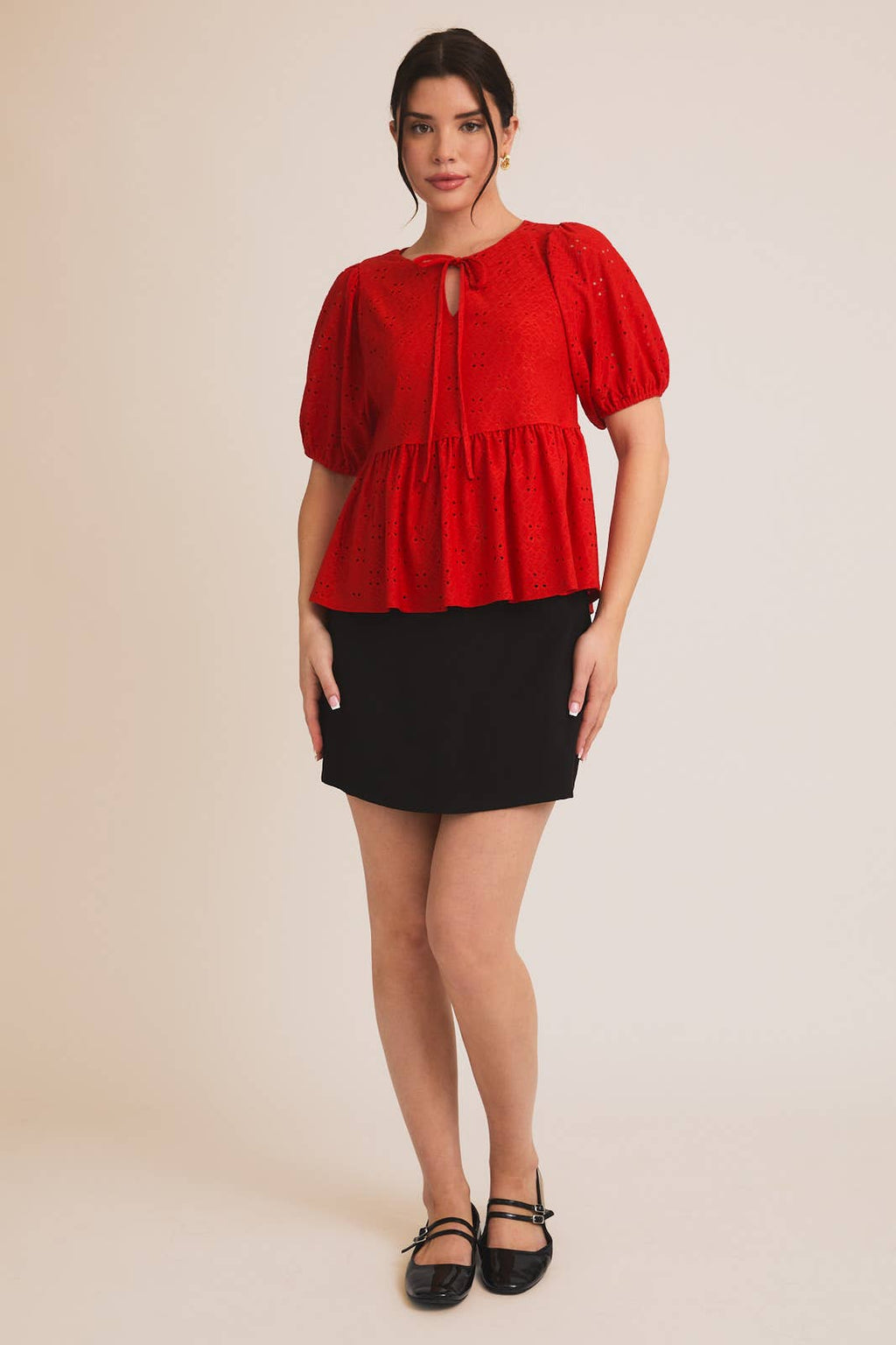 Woman wearing a red eyelet babydoll  top with puff sleeves and a black skirt on a beige background