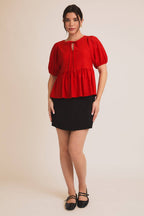 Woman wearing a red eyelet babydoll  top with puff sleeves and a black skirt on a beige background