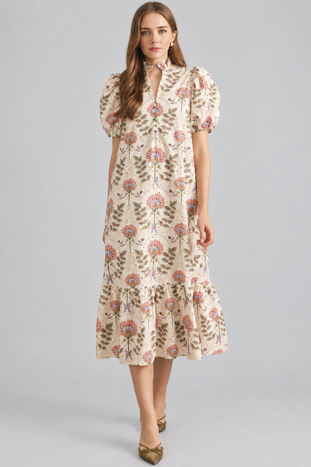 A woman modeling a cream maxi dress with brown, pink, and green floral pattern on eyelet fabric, standing against a light beige brick wall.