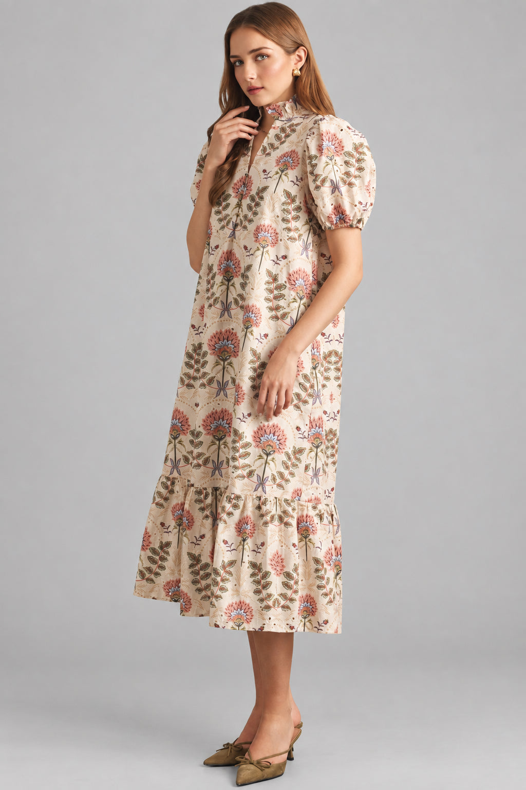 A woman modeling a cream maxi dress with brown, pink, and green floral pattern on eyelet fabric, standing against a light beige brick wall.