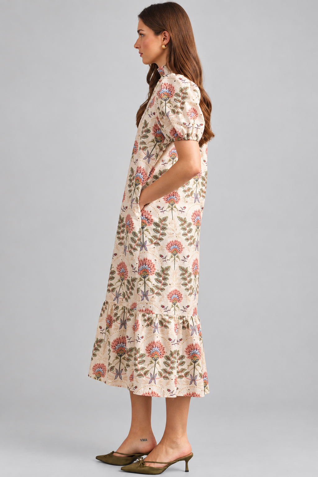 A woman modeling a cream maxi dress with brown, pink, and green floral pattern on eyelet fabric, standing against a light beige brick wall.