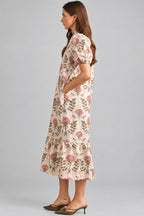 A woman modeling a cream maxi dress with brown, pink, and green floral pattern on eyelet fabric, standing against a light beige brick wall.