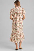 Back view of a woman modeling a cream maxi dress with brown, pink, and green floral pattern on eyelet fabric, standing against a light beige brick wall.