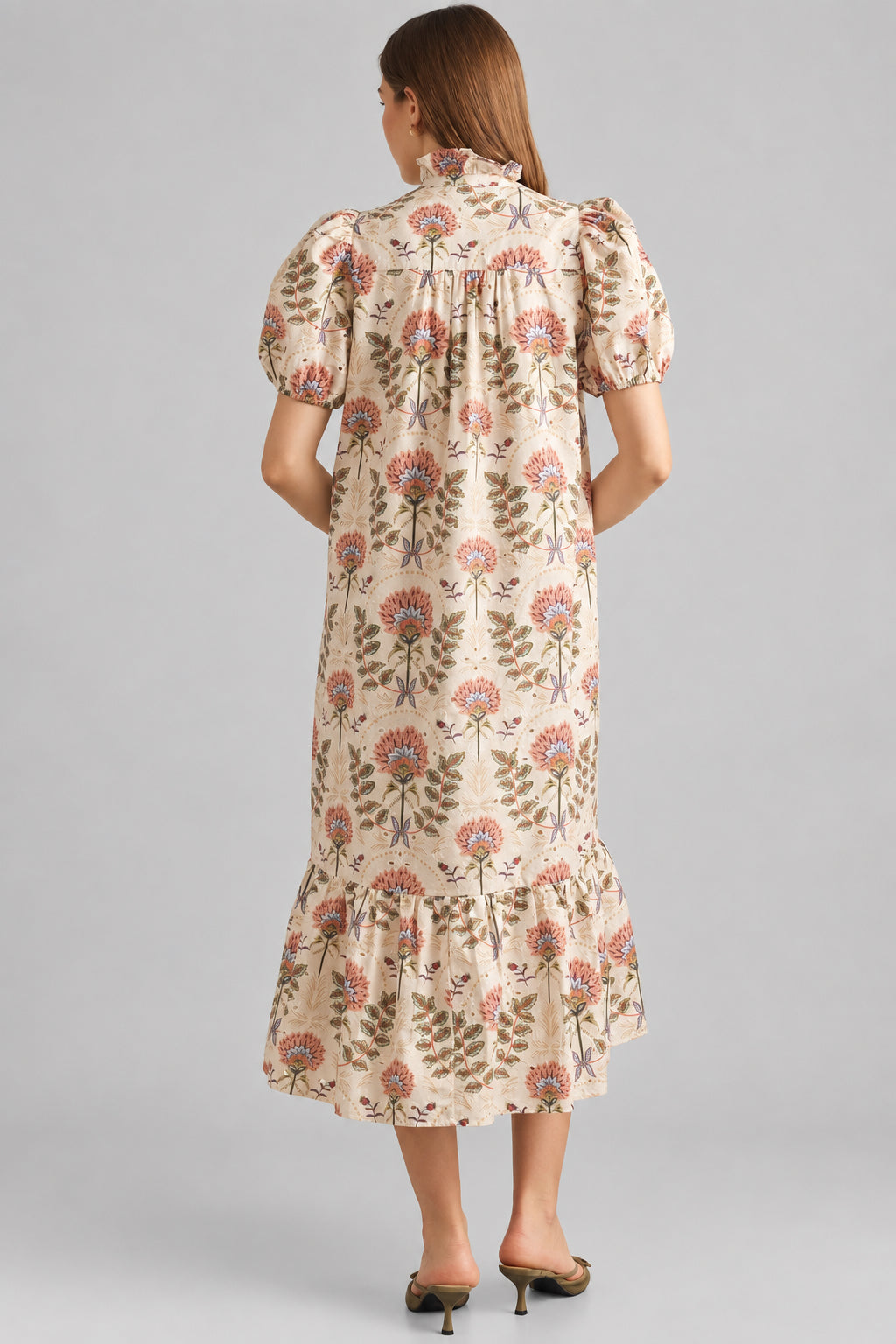 Back view of a woman modeling a cream maxi dress with brown, pink, and green floral pattern on eyelet fabric, standing against a light beige brick wall.