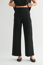 A model wearing black wide-leg cropped pants, paired with a black crop top and black flat shoes, shown from the waist down against a light gray studio background.