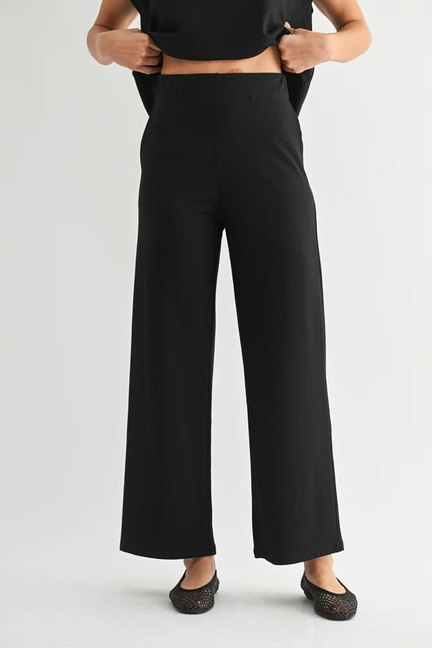 A model wearing black wide-leg cropped pants, paired with a black crop top and black flat shoes, shown from the waist down against a light gray studio background.