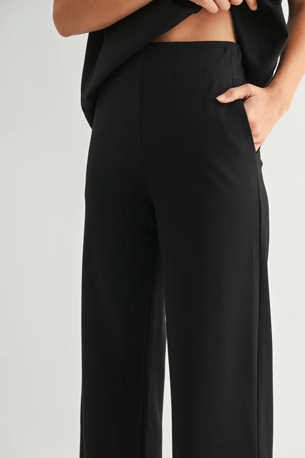 Close-up of a model wearing black wide-leg trousers, with one hand in the pocket and the other lifting a black top, against a light gray studio background.