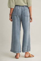 Back view of a woman wearing cropped drawstring medium blue denim wide-leg pants on a beige background