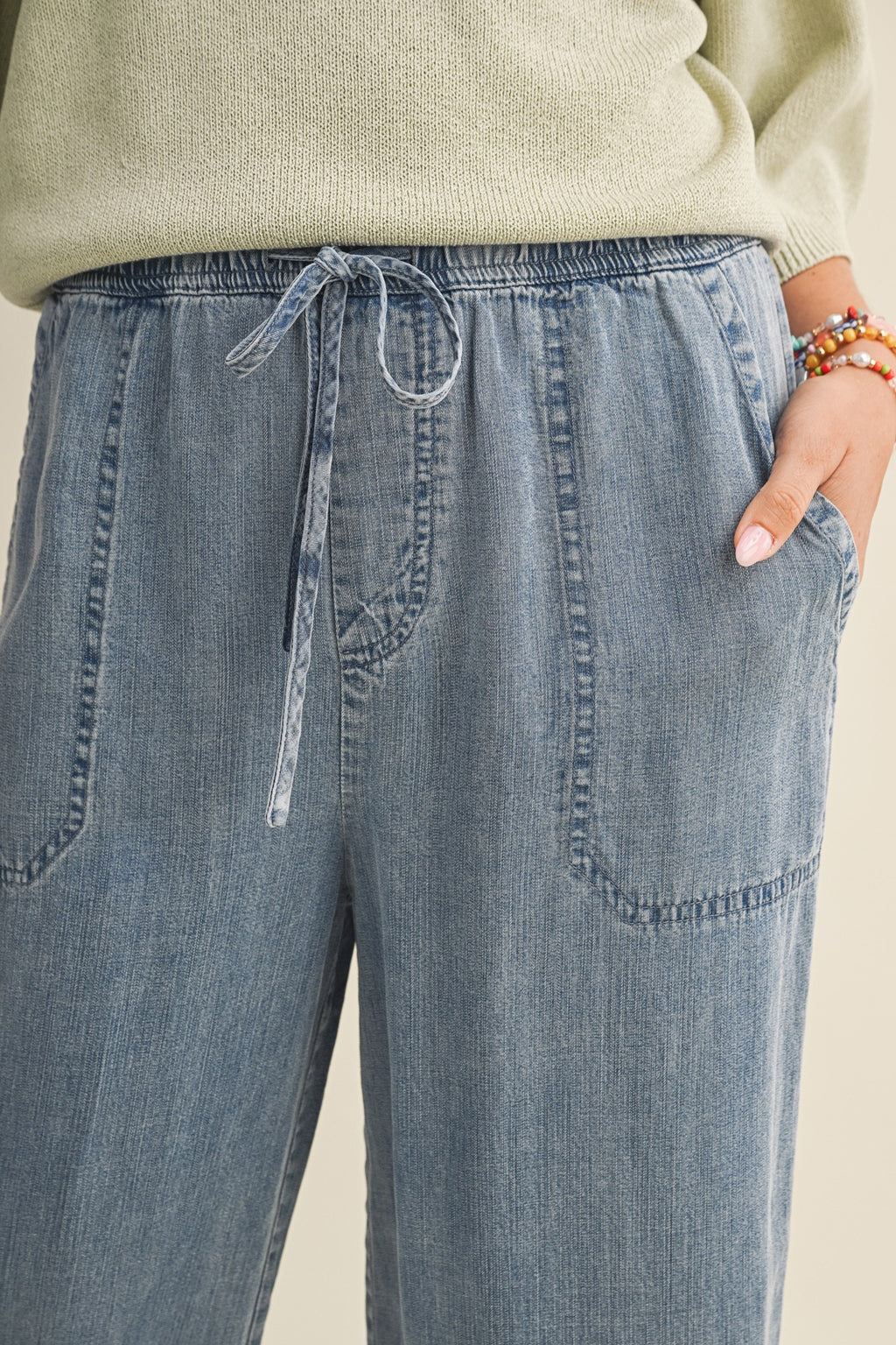 Close up of a woman wearing cropped drawstring medium blue denim wide-leg pants on a beige background
