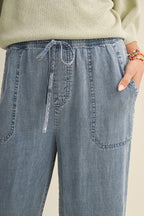 Close up of a woman wearing cropped drawstring medium blue denim wide-leg pants on a beige background
