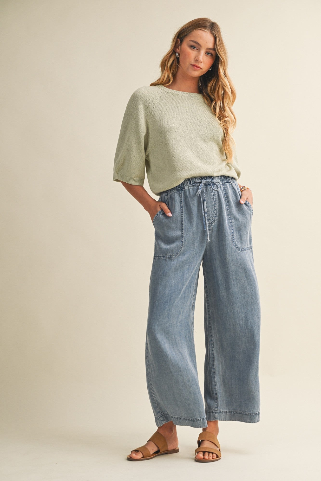 Woman wearing cropped drawstring medium blue denim wide-leg pants with gray top on a beige background