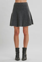 A woman wearing a gray flared knit mini skirt and black leather ankle boots, standing against a solid light beige studio background.