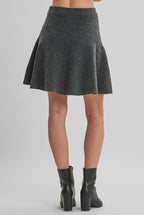 Back view of a  woman wearing a gray flared knit mini skirt and black leather ankle boots, standing against a solid light beige studio background.