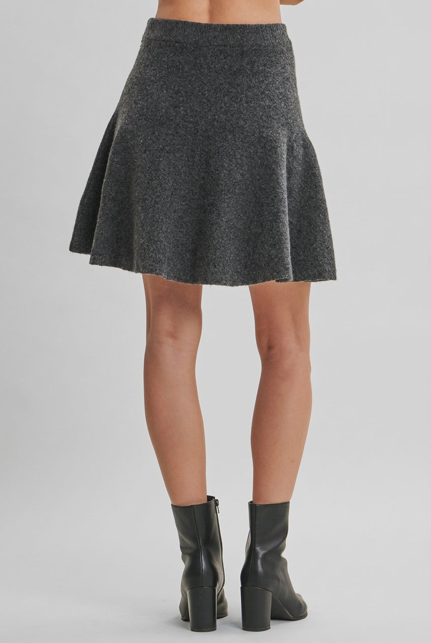Back view of a  woman wearing a gray flared knit mini skirt and black leather ankle boots, standing against a solid light beige studio background.