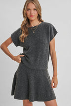 A woman wearing a gray short sleeve sweater and flared knit mini skirt standing against a solid light beige studio background.