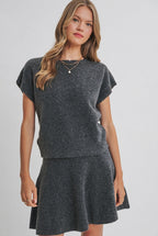A woman wearing a gray short sleeve sweater and flared knit mini skirt standing against a solid light beige studio background.