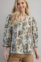 A woman wearing a light sage green floral blouse with brown and rust flowers, paired with tan pants, against a solid gray studio background.