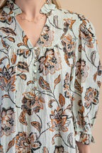 Close up of a light sage green floral blouse with brown and rust flowers, paired with tan pants, against a solid gray studio background.