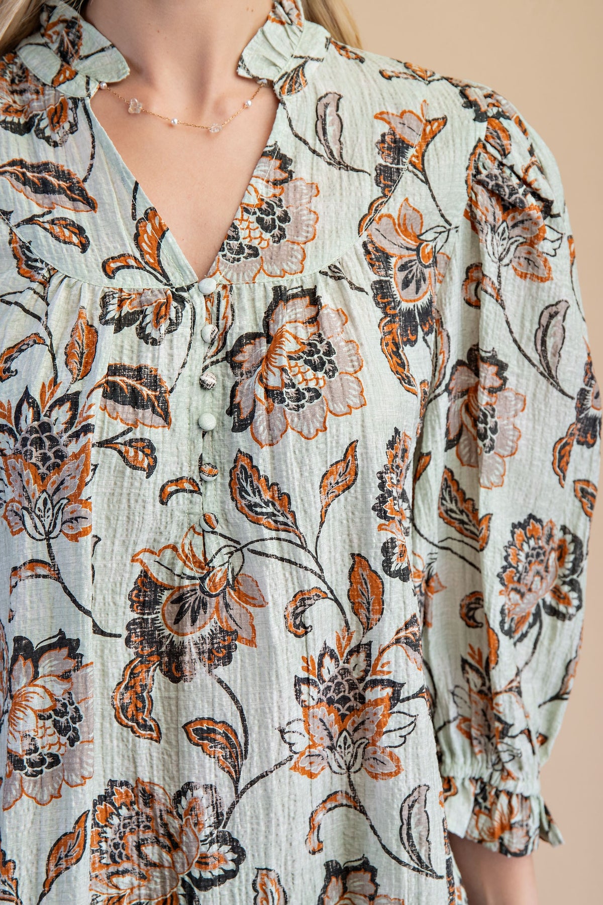 Close up of a light sage green floral blouse with brown and rust flowers, paired with tan pants, against a solid gray studio background.