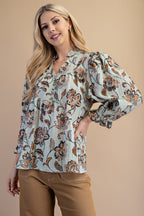 A woman wearing a light sage green floral blouse with brown and rust flowers, paired with tan pants, against a solid gray studio background.