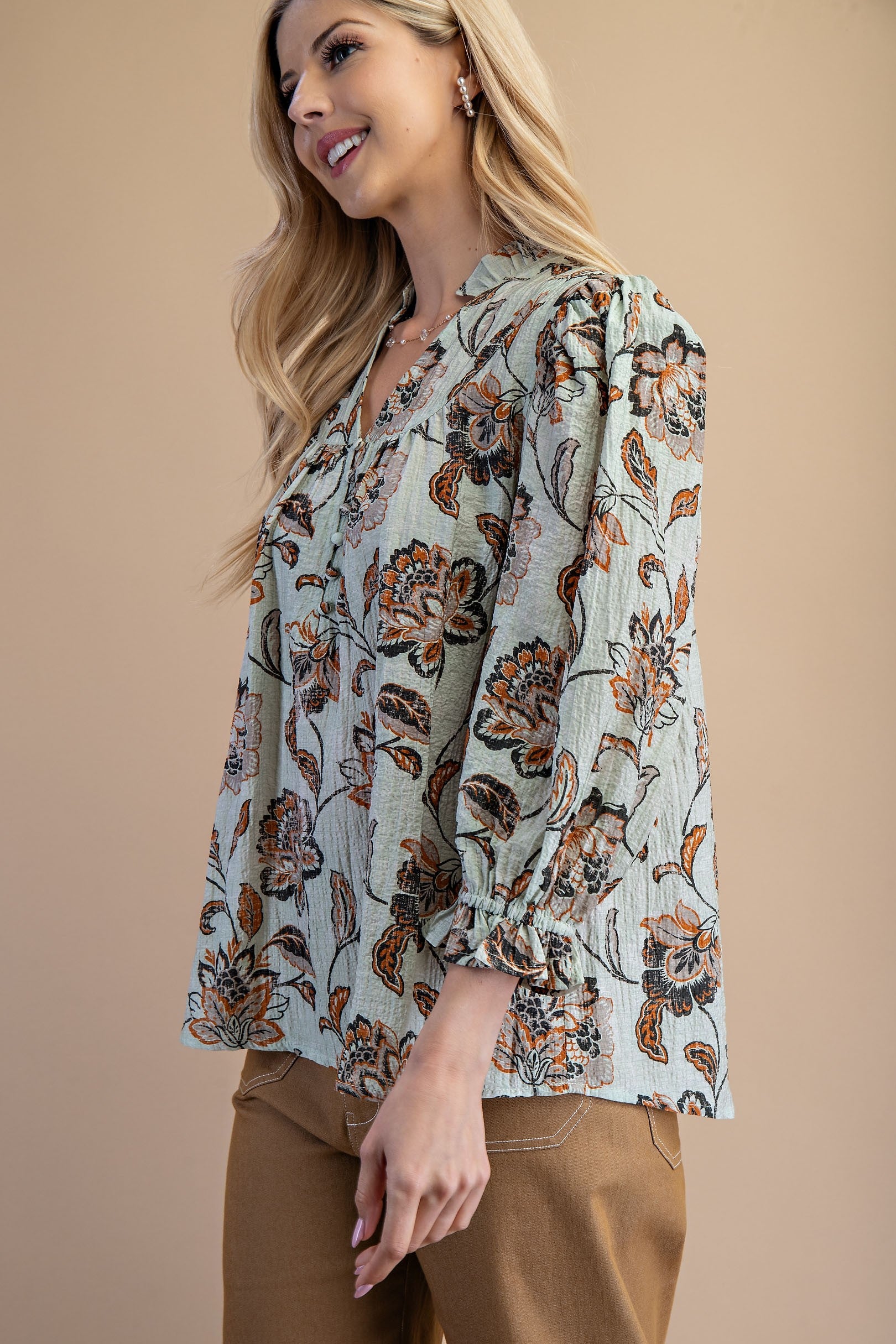 A woman wearing a light sage green floral blouse with brown and rust flowers, paired with tan pants, against a solid gray studio background.