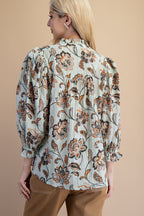 Back view of a woman wearing a light sage green floral blouse with brown and rust flowers, paired with tan pants, against a solid gray studio background.