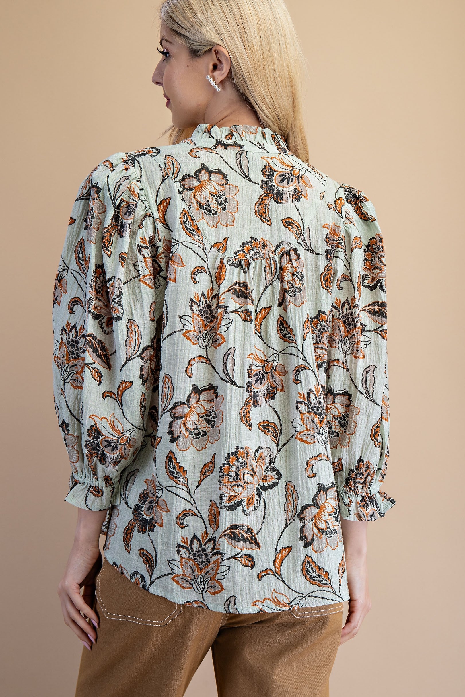 Back view of a woman wearing a light sage green floral blouse with brown and rust flowers, paired with tan pants, against a solid gray studio background.
