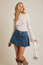 Woman wearing a oatmeal ribbed knit  top and blue denim skirt holding a beige bag against a neutral background