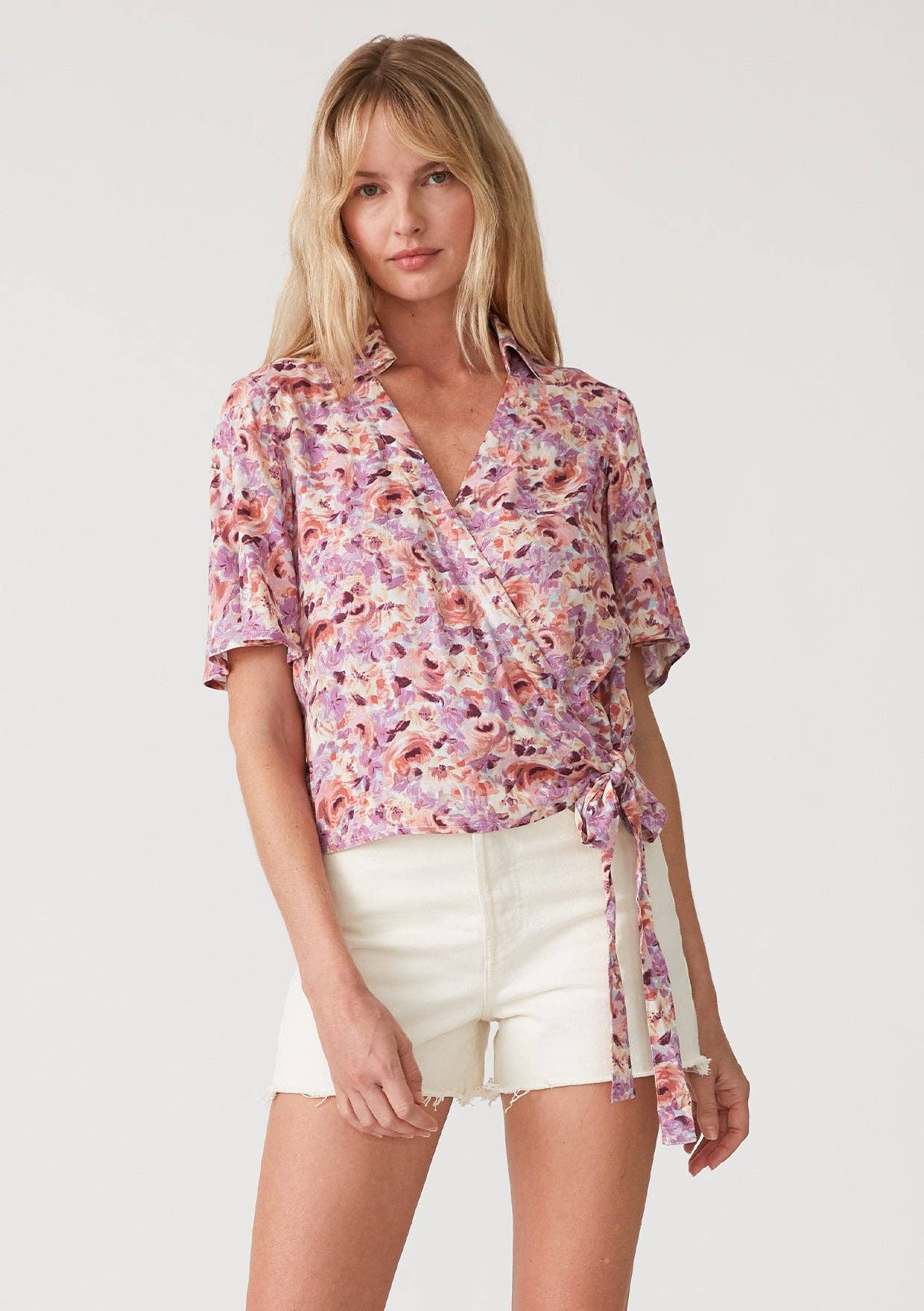 Woman wearing a floral wrap top with side tie and white shorts on a plain background