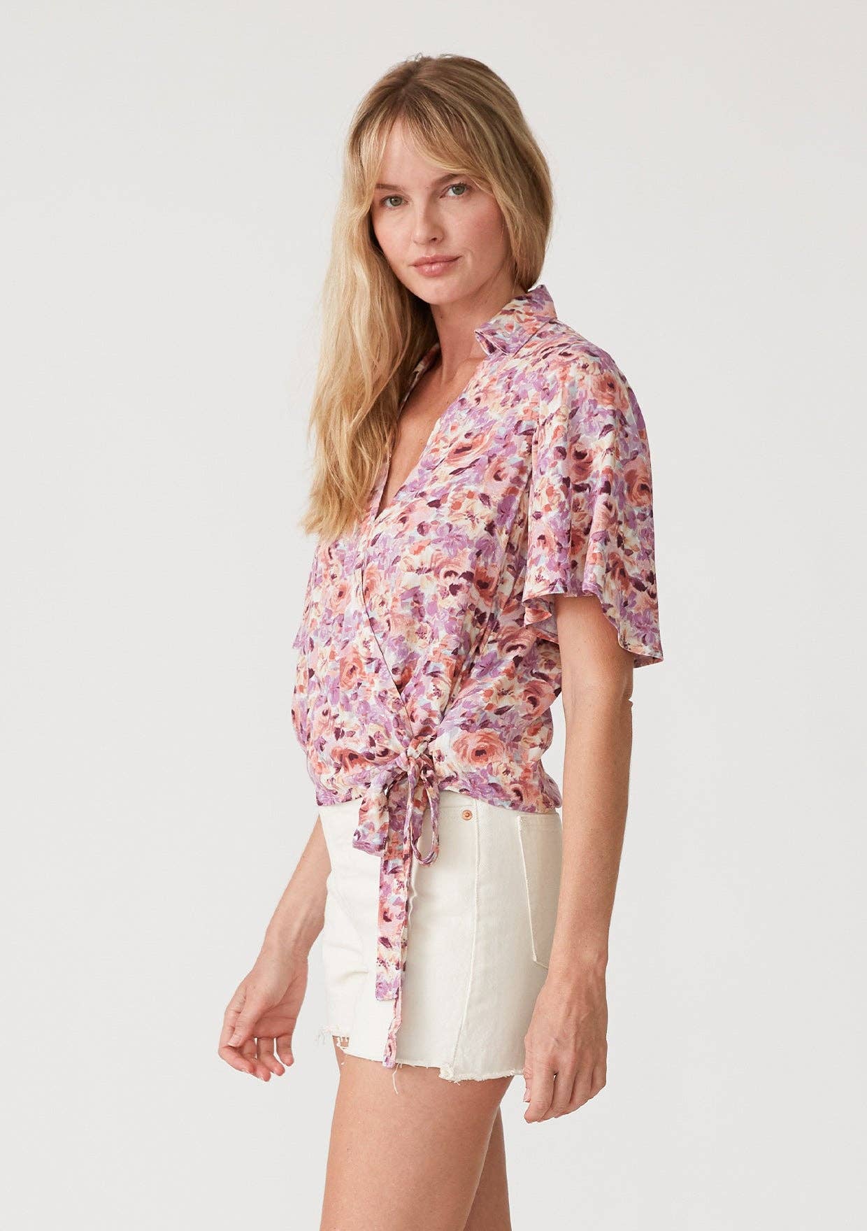 Woman wearing a floral wrap top with side tie and white shorts on a plain background